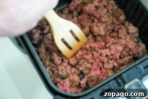 Ground beef sizzling and browning in the air fryer basket, being stirred with a wooden spatula.