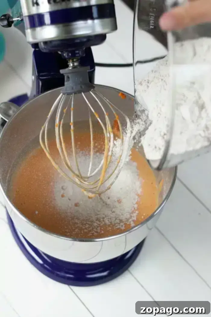 Half Sheet Cake Recipe 3 A vibrant orange cake batter being mixed in a stand mixer, with a whisk attachment visible and flour being added from a glass bowl.