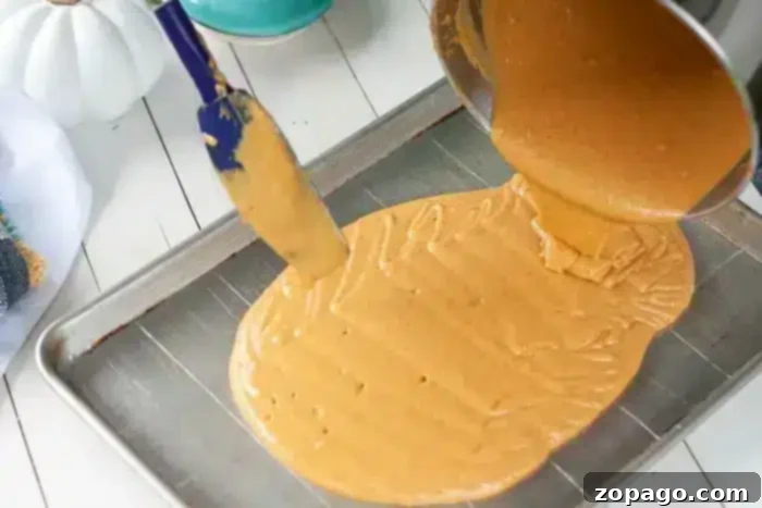 Half Sheet Cake Recipe 4 A rich, caramel-colored cake batter is being carefully poured from a mixing pot into a large metal baking sheet, prepared for baking a half sheet cake.