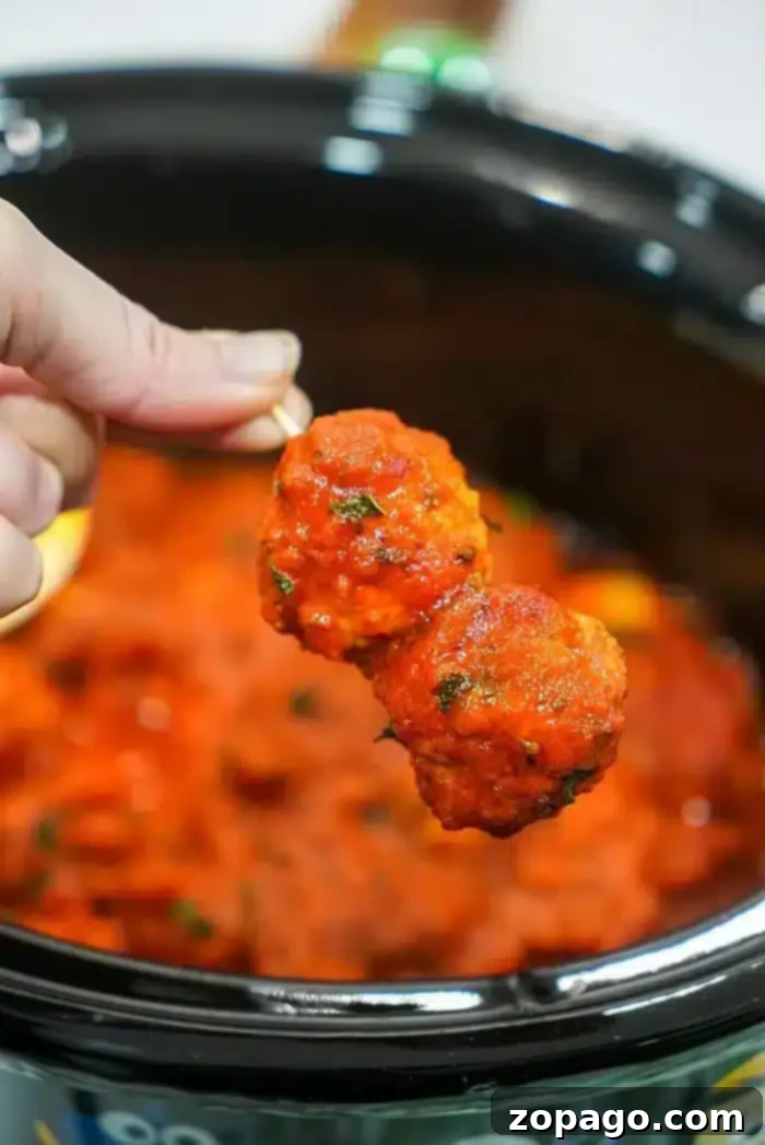 Effortless Crockpot Frozen Meatballs 2 A hand holds a skewer with two succulent meatballs coated in tomato sauce, poised above a crockpot filled with more simmering meatballs and sauce. Using frozen meatballs in the crockpot makes this dish an effortless delight.