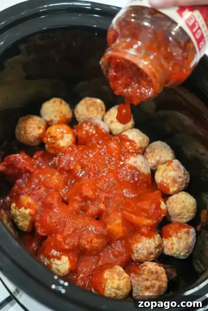 Effortless Crockpot Frozen Meatballs 6 Frozen meatballs in the crockpot are being covered with tomato sauce poured from a jar.