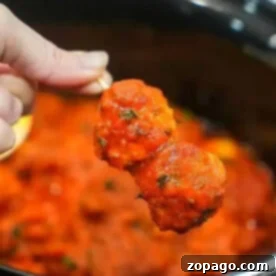 Effortless Crockpot Frozen Meatballs 8 A hand holds two frozen meatballs covered in spaghetti sauce over a pot filled with more sauce, ready to simmer perfectly in the crockpot.