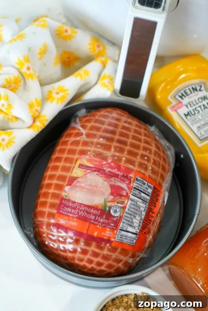 Crispy Air Fryer Ham 3 A round pan containing a packaged hickory-smoked cooked whole ham sits on a counter, ready to be transformed into a delicious air fryer ham. Surrounding items include a tub of yellow mustard, a small container of brown sugar, a meat thermometer, and a cloth with a yellow floral pattern.