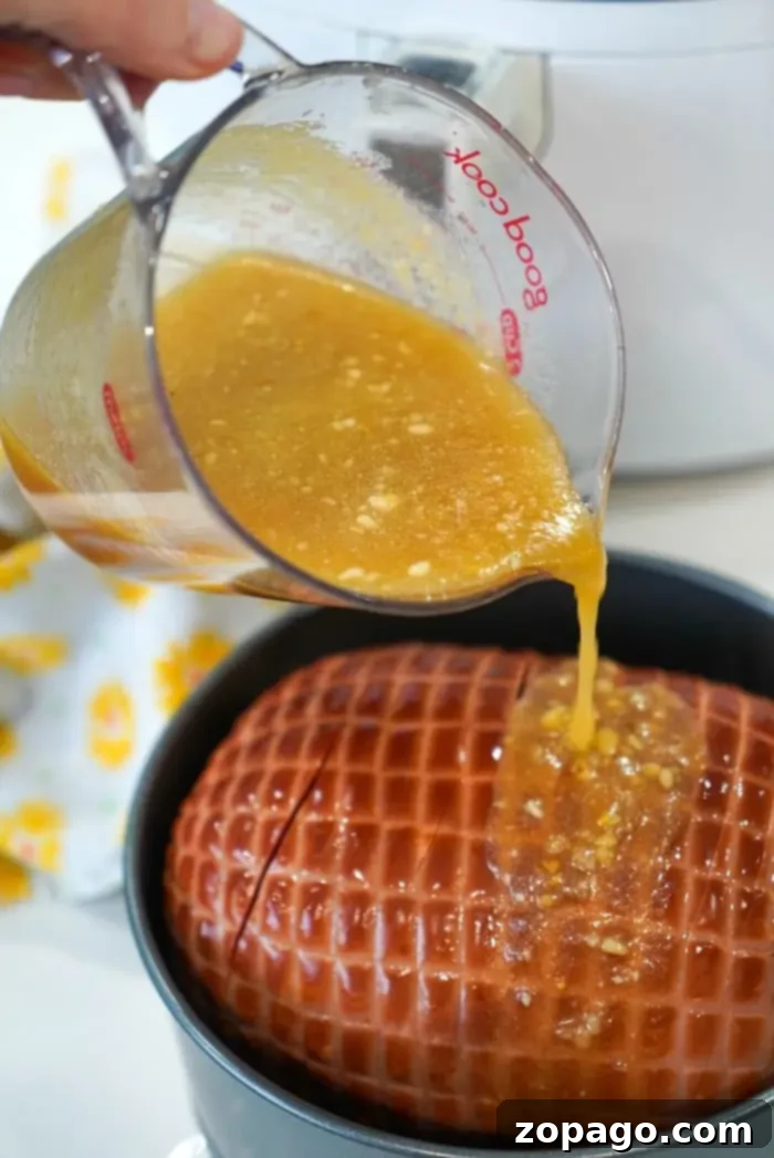 Perfect Air Fryer Ham 4 A hand is gently pouring a luscious pineapple glaze from a glass measuring cup over a scored air fryer ham, nestled in a black cooking pot.