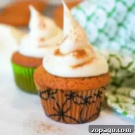 Two gingerbread muffins topped with a swirl of cream and a sprinkle of cinnamon sit on a white surface. The muffins are in decorative wrappers, one green and one with a black pattern. A green and white patterned cloth is partially visible in the background.
