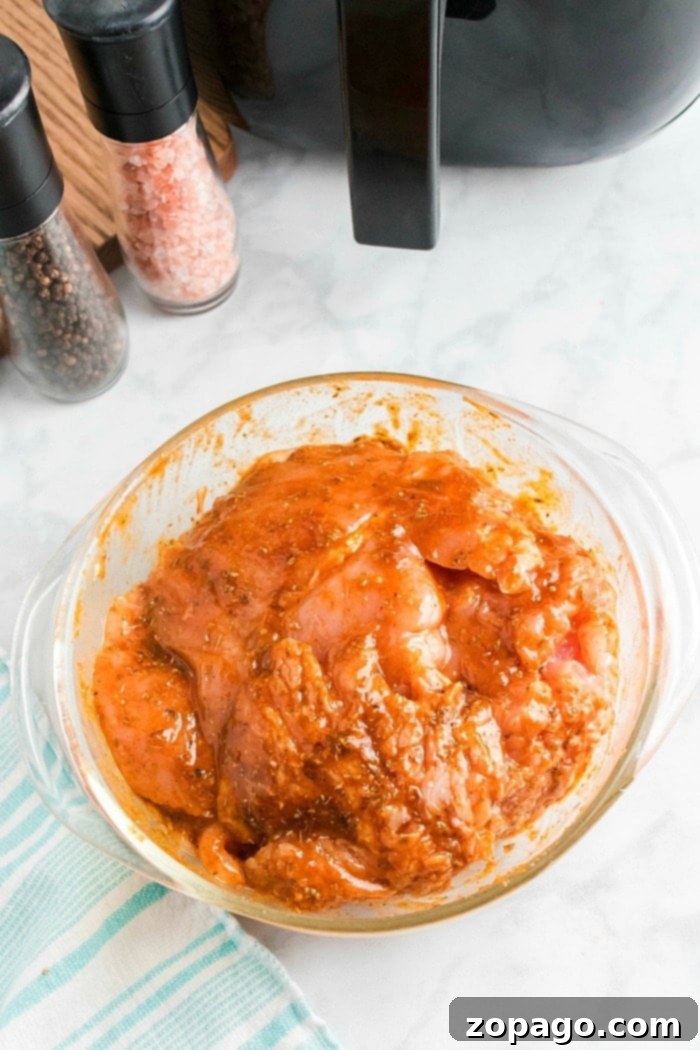 Crispy Air-Fried Turkey Breast 5 A glass bowl filled with raw boneless turkey breast marinating in a reddish sauce is placed on a marble countertop. Nearby, an air fryer hints at future meals, alongside salt and pepper grinders, a striped cloth, and a black appliance.