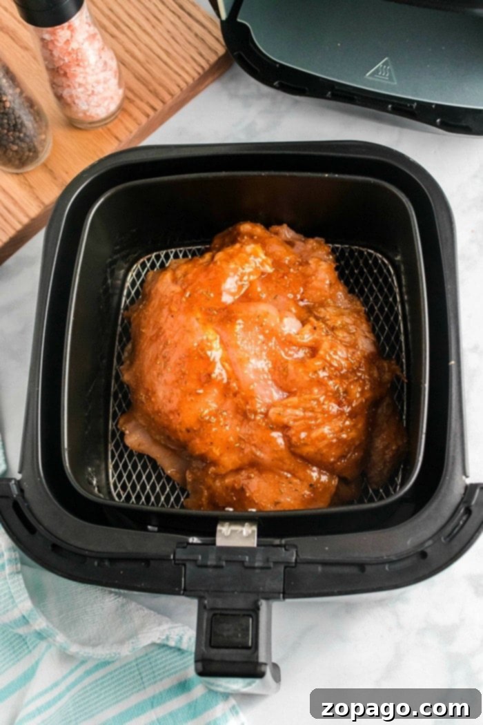 Crispy Air-Fried Turkey Breast 6 Raw marinated turkey breast placed in an air fryer basket, ready to cook alongside the succulent aroma of air fryer turkey breast. The air fryer lid is open, revealing its contents. A wooden board with salt and pepper shakers is visible in the background, with a blue and white cloth partially seen at the bottom.