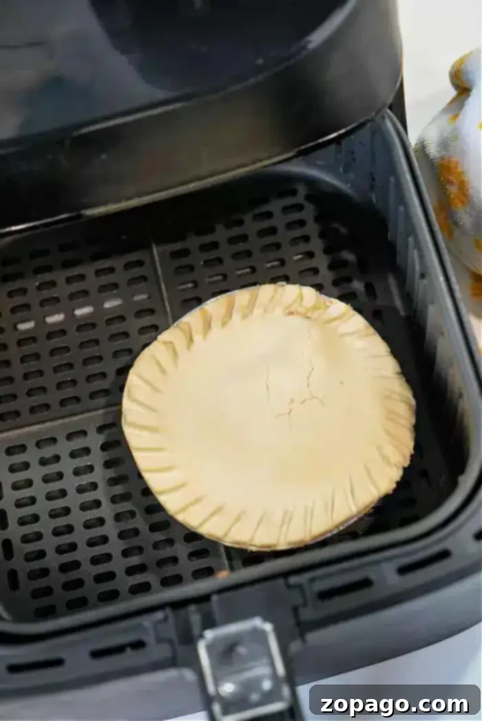 Crispy Air Fryer Frozen Pot Pie 5 A frozen pot pie with a crimped edge and cracked top sits in the basket of an air fryer. The interior surface, complete with black perforated grates, is visible. On the side, a partial view of a patterned cloth adds a touch of charm.