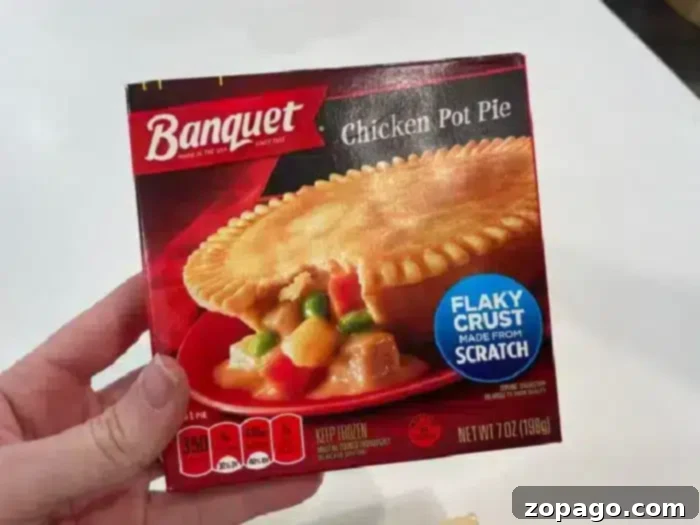Crispy Air Fryer Frozen Pot Pie 7 A person's hand is holding a box of Banquet Chicken Pot Pie. The packaging showcases a flaky crust, made from scratch, and suggests cooking the 7-ounce frozen pot pie in an air fryer for optimal crispiness. Nutritional info and instructions are detailed on the box.