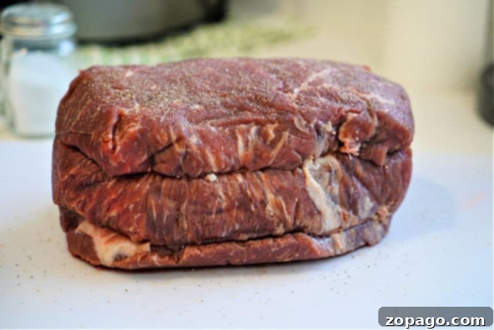 Slow-Cooked Dutch Oven Roast 3 A raw beef roast sits on a white cutting board, lightly seasoned with salt and pepper, ready to become a tender Dutch oven roast. A salt shaker and a green and white cloth are blurred in the background.
