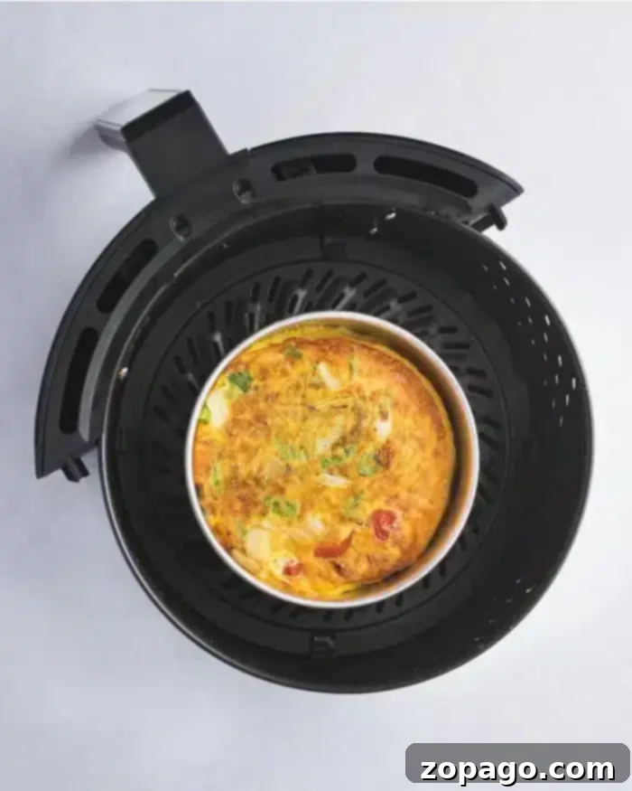 Air Fryer Quiche Perfection 8 An air fryer with a removable basket contains a delicious quiche, featuring visible ingredients like tomatoes, green herbs, and cheese. The appliance is placed on a light-colored surface.