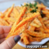 Close-up of a hand holding crispy air fryer carrot fries, with a plate of these deliciously baked carrot fries in the background.
