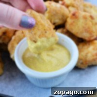 Homemade Crispy Air Fryer Chicken Nuggets 6 A hand dips a crispy homemade chicken nugget, fresh from the air fryer, into a small bowl of mustard sauce on a tray brimming with nuggets.