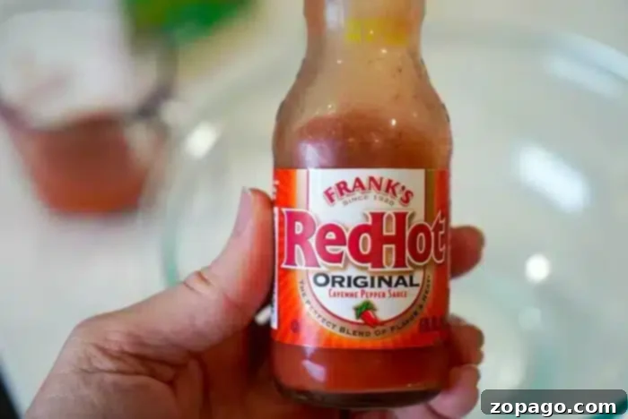 Copycat Bang Bang Shrimp Sauce 4 A hand holds a bottle of Frank's RedHot Original Cayenne Pepper Sauce with its signature red label. In the blurry background, a measuring cup and glass bowl hint at a delicious creation—perhaps the perfect bang bang shrimp sauce in the making.