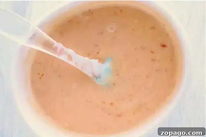 Copycat Bang Bang Shrimp Sauce 5 A plastic spoon is dipped into a container of creamy bang bang shrimp sauce, with visible flecks of seasoning throughout. The sauce has a light orange hue and a smooth texture, reminiscent of Thousand Island dressing.