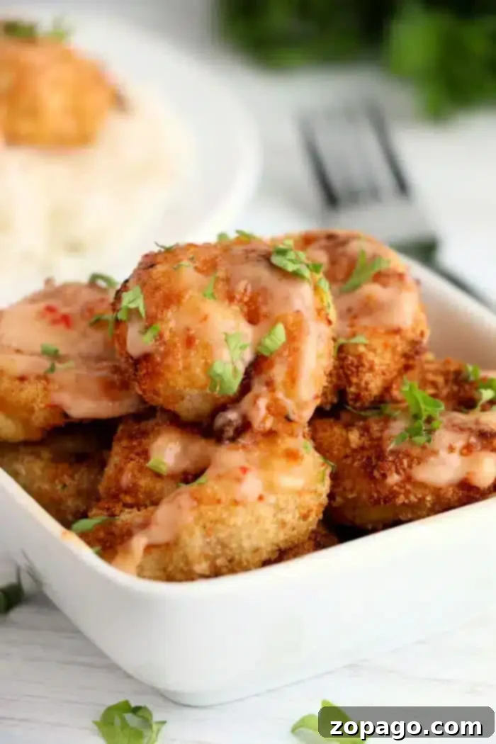 Copycat Bang Bang Shrimp Sauce 6 A rectangular white dish filled with crispy fried shrimp drizzled with bang bang shrimp sauce. The shrimp are garnished with chopped parsley. A fork and a plate of rice are partially visible in the background.