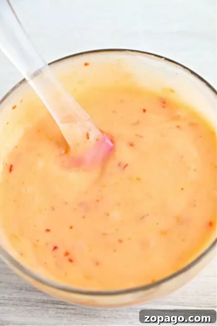 Copycat Bang Bang Shrimp Sauce 8 A glass bowl filled with a creamy, orange bang bang shrimp sauce spotted with small red specks. A clear spoon rests inside the bowl, ready to serve. The surface beneath is a light, textured material.