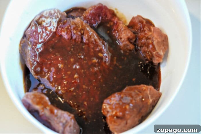 Easy Air Fryer Cube Steak 7 Raw cube steak pieces marinating in a dark, thick sauce in a white bowl, perfect for an air fryer feast.