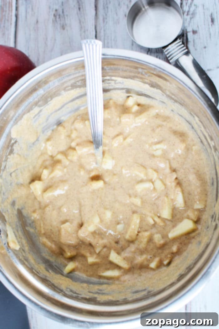 Air Fryer Apple Fritter Perfection 5 A bowl of fresh apple fritter batter with a spoon, beside a red apple and measuring cup on a wooden surface.