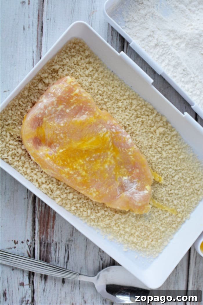 Raw chicken cutlet coated in egg on a tray of breadcrumbs, ready to become delicious chicken cutlets in the air fryer, with a spoon nearby.