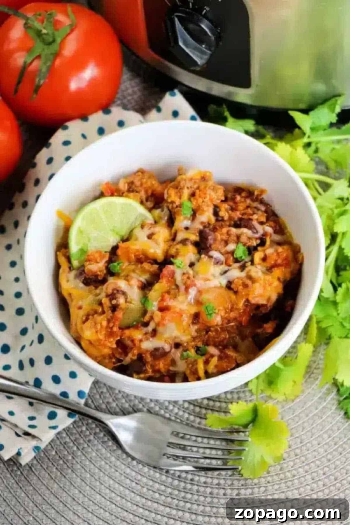 Easy Slow Cooker Enchilada Casserole 2 A steaming bowl of cheesy Crockpot enchilada casserole, richly topped with melted cheese, a fresh lime wedge, and garnished with cilantro, served alongside ripe tomatoes and a fork.