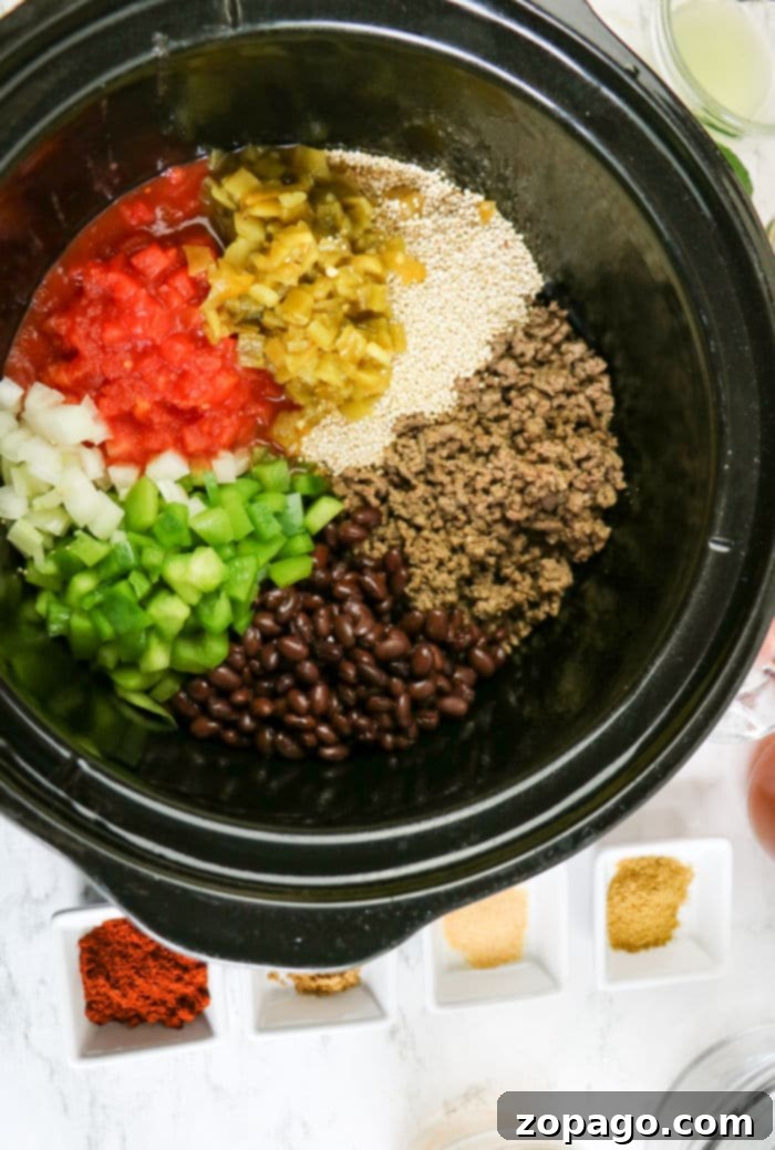 Easy Slow Cooker Enchilada Casserole 4 Top-down view of a Crockpot filled with colorful ingredients for enchilada casserole: dark black beans, browned ground beef, bright diced tomatoes, green peppers, white onions, golden quinoa, and various spices, all ready to cook.