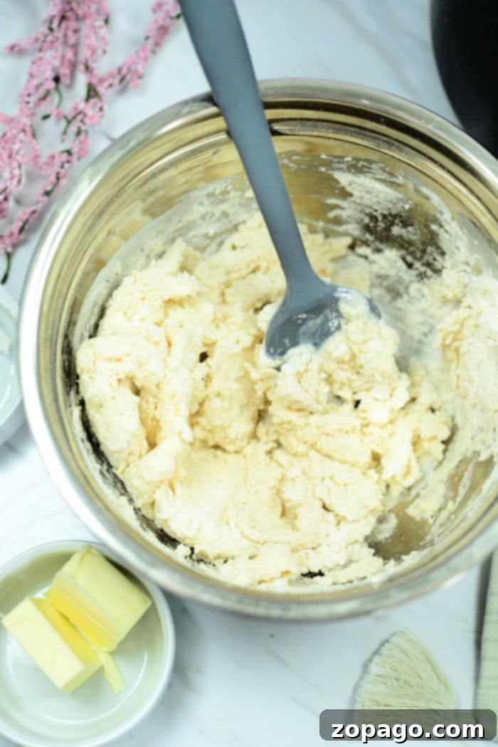 Golden Air Fryer Biscuits 3 A metal mixing bowl contains a dough mixture with a grey spatula resting inside, perfect for crafting air fryer biscuits. Nearby, a small dish holds two pieces of butter. A few pink flowers are partially visible in the background on a white surface.