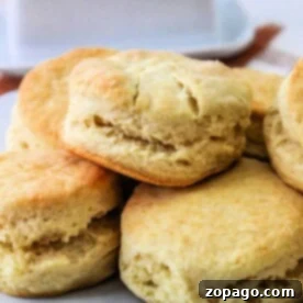 A plate of freshly baked air fryer biscuits stacked on top of each other. The golden brown biscuits boast a soft, fluffy texture. A butter dish is blurred in the background.