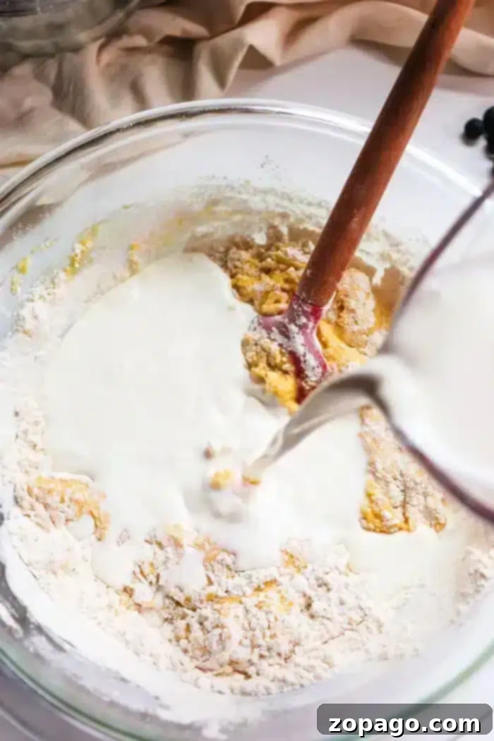 Just the Tops: Blueberry Muffin Perfection 5 A glass mixing bowl holds flour and egg batter being combined with a wooden spoon. A pitcher is actively pouring liquid, likely milk, into the bowl, preparing the mixture for delightful blueberry muffin tops. The scene is set against a beige cloth with scattered blueberries.