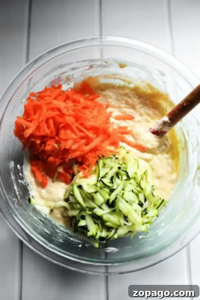 Zucchini Carrot Morning Glory Muffins 4 A glass bowl contains batter mixed with grated carrots and zucchini, perfect for making delicious zucchini carrot muffins. A wooden spoon is partially submerged in the mixture, resting on a white countertop.