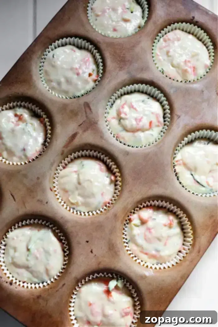 Zucchini Carrot Morning Glory Muffins 6 A muffin tin holds eleven cupcake liners filled with zucchini carrot muffins batter, showcasing pieces of red and green ingredients. The tin rests on a white surface.