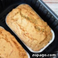 Two loaves of golden-brown zucchini bread in metal trays rest perfectly inside a sleek black air fryer basket.
