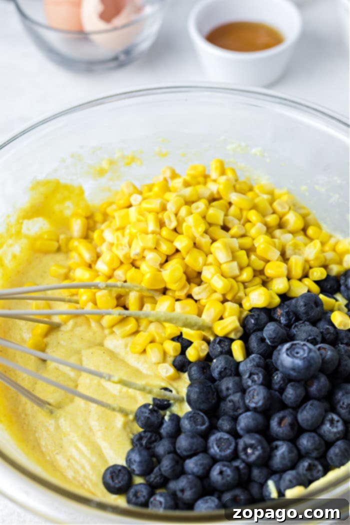 Homestyle Cast Iron Cornbread 6 A glass bowl holds cornmeal batter adorned with corn and blueberries, with a whisk at the ready; eggshells and a cup linger in the background, setting the stage for delicious Jiffy cornbread baked to perfection in a cast iron skillet.