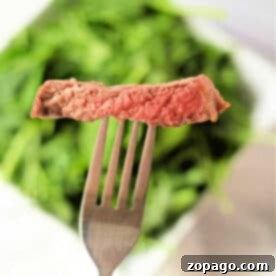 A fork holds a slice of rare ribeye in air fryer steak above a blurred background of green leafy vegetables.