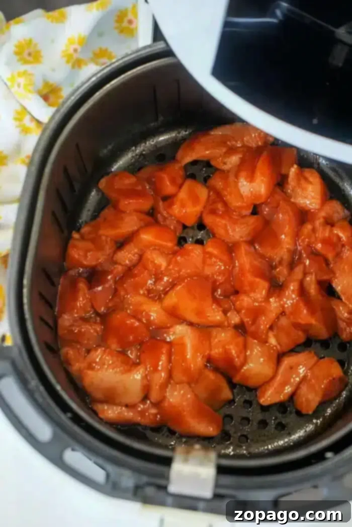 Juicy Air Fryer BBQ Chicken Breast 10 Chunks of marinated chicken are placed inside an open air fryer basket, sizzling into succulent air fryer BBQ chicken. A white kitchen towel with yellow floral patterns is partially visible on the side.
