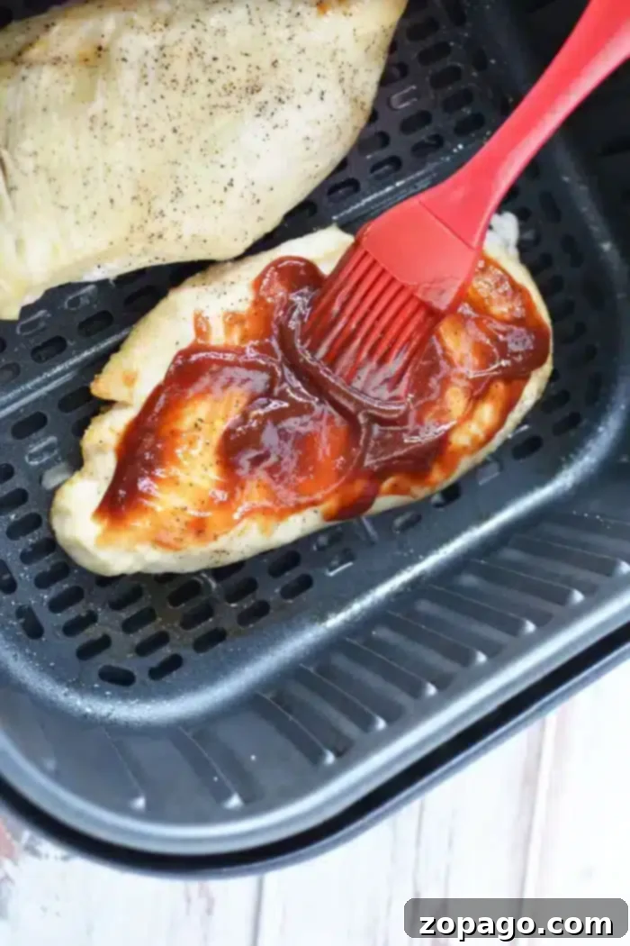 Juicy Air Fryer BBQ Chicken Breast 7 A piece of air fryer BBQ chicken is being brushed with barbecue sauce using a red silicone brush inside the basket. Another piece of seasoned chicken sits tantalizingly in the background, ready to be savored.