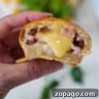 A hand holds a baked bread roll reminiscent of air fryer chicken cordon bleu, filled with melted cheese and small pieces of ham. A bite reveals the gooey interior, while greenery and other ingredients blur in the background.