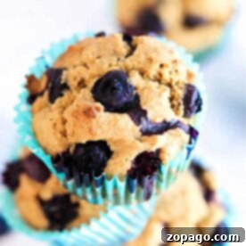 A blueberry bran muffin with a golden crust sits in a turquoise wrapper against a blurred background.
