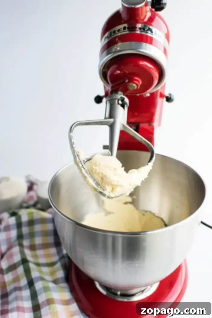 Effortless 3-Ingredient Shortbread 4 A vibrant red stand mixer busily whips a creamy mixture in a metal bowl, using a paddle attachment. The smooth, consistent texture of the mixture is clearly visible, set against a simple white wall backdrop with a plaid cloth partially seen on the left.