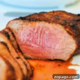 Close-up of a cooked air fryer tri-tip steak, sliced to reveal a pink, juicy interior. The steak boasts a seasoned, grilled crust with some juices pooled around it on the white plate.
