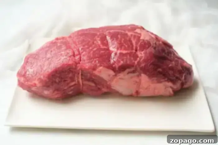 Ultimate Air Fryer Roast Beef 3 A prime raw beef roast, showing beautiful marbling and a rich red color, resting on a clean white platter, ready for seasoning and air frying. The background is softly blurred to emphasize the quality of the meat.