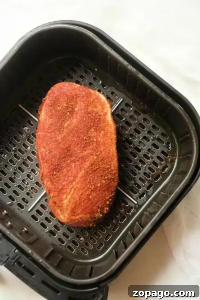 A piece of seasoned pork roast, ready for cooking, sits in a black air fryer basket with its perforated design for optimal air circulation, highlighting the applied reddish spice mix.