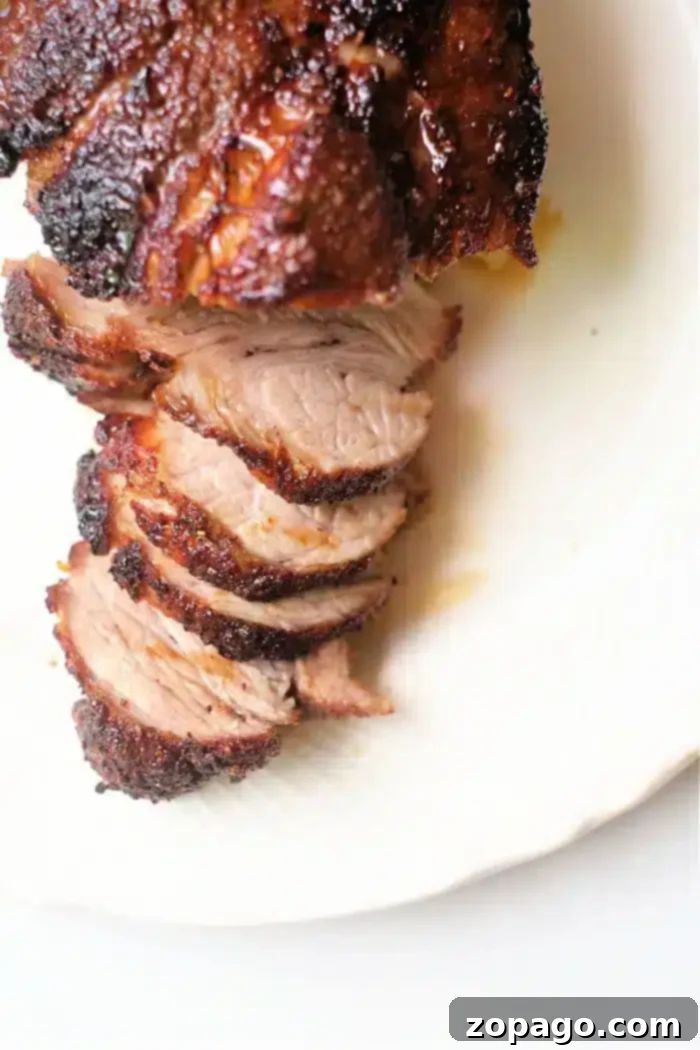 Crispy Air Fryer Pork Roast 5 A close-up view of a roasted, sliced air fryer pork roast on a white plate. The meat is browned and crispy on the outside, with a juicy, tender interior. Five slices are visible, arranged neatly in a row.