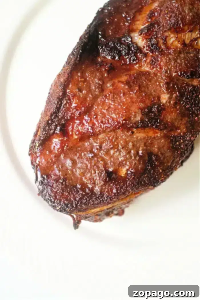 A close-up image of a well-cooked pork roast, showcasing a crispy, seared crust and juicy, tender texture on a white plate, seasoned with flavorful spices and herbs.