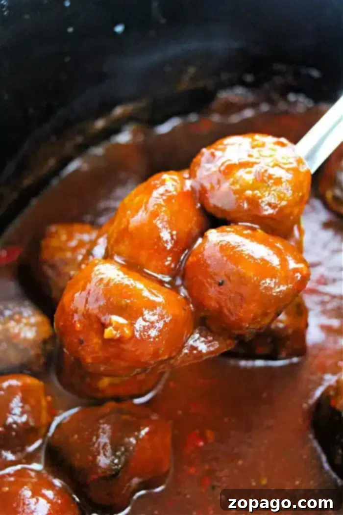 Grape Chili Glazed Meatballs 2 A spoon lifts several glazed meatballs, perfectly cooked as a crockpot appetizer, from a pot filled with thick, brown sauce. The glossy surface of the sauce clings to the meatballs, suggesting they are beautifully marinated and ready to delight at any gathering.
