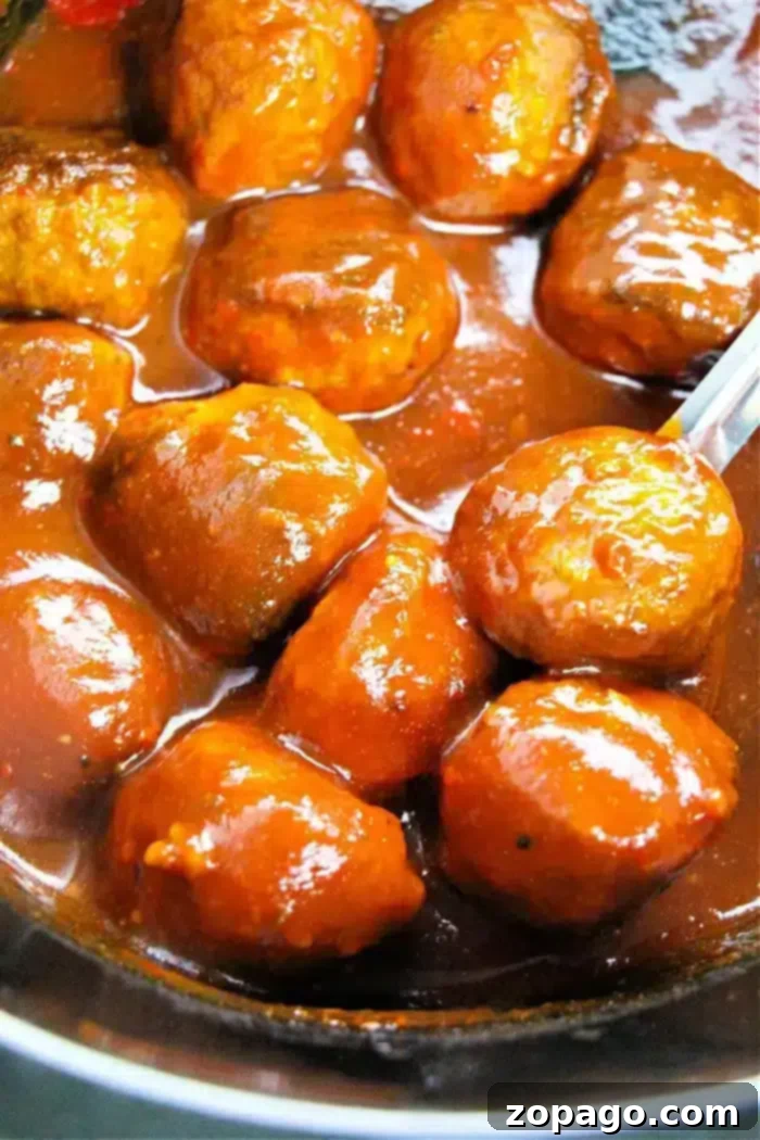 Grape Chili Glazed Meatballs 3 Close-up of several savory meatballs covered in a rich, glossy sauce in a pan. A spoon is partially visible on the right, suggesting stirring or serving. The sauce appears thick and evenly coats the meatballs, reminiscent of crockpot appetizer meatballs with grape jelly.