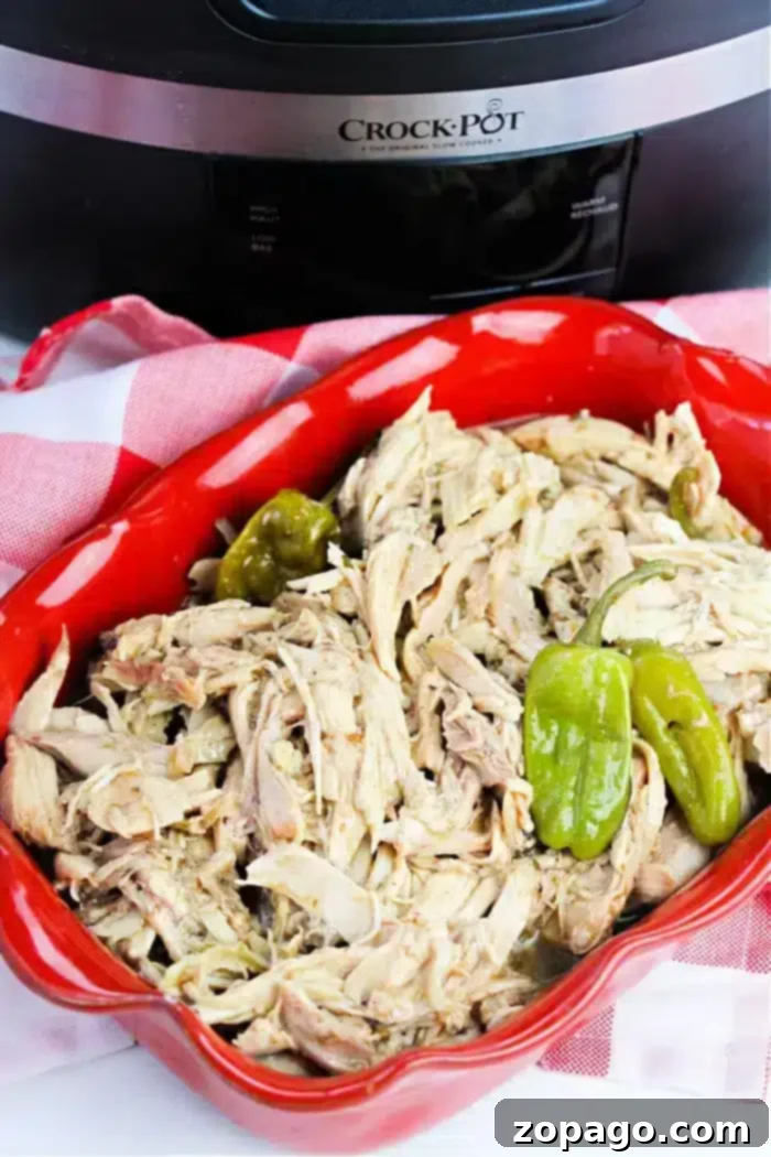 Zesty Pepperoncini Chicken 2 A vibrant red dish filled with succulent shredded pepperoncini chicken and whole green peppers, artfully arranged on a classic white and red checkered cloth. In the soft-focus background, a black Crock-Pot slow cooker hints at the ease of preparation.