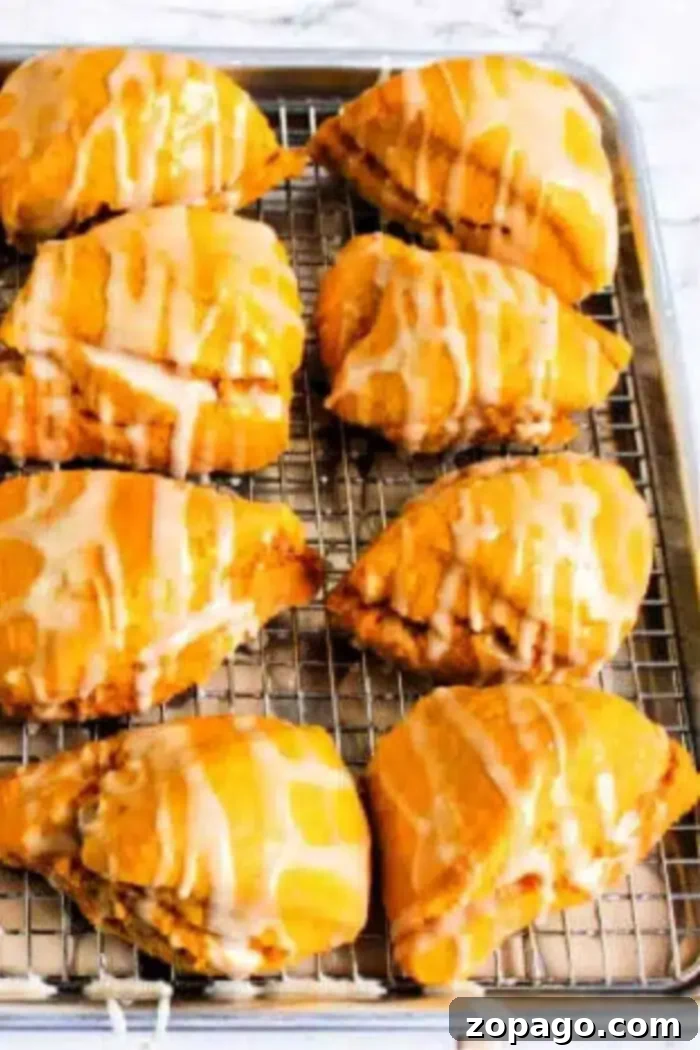 Eight golden brown pastries, reminiscent of a Starbucks pumpkin scone, with a creamy glaze drizzle, are arranged neatly on a cooling rack placed over a baking sheet, ready to be enjoyed.