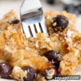 Close-up of a fork piercing a slice of blueberry bread pudding, its crumb-topped layers offering a delectable treat on a white plate.