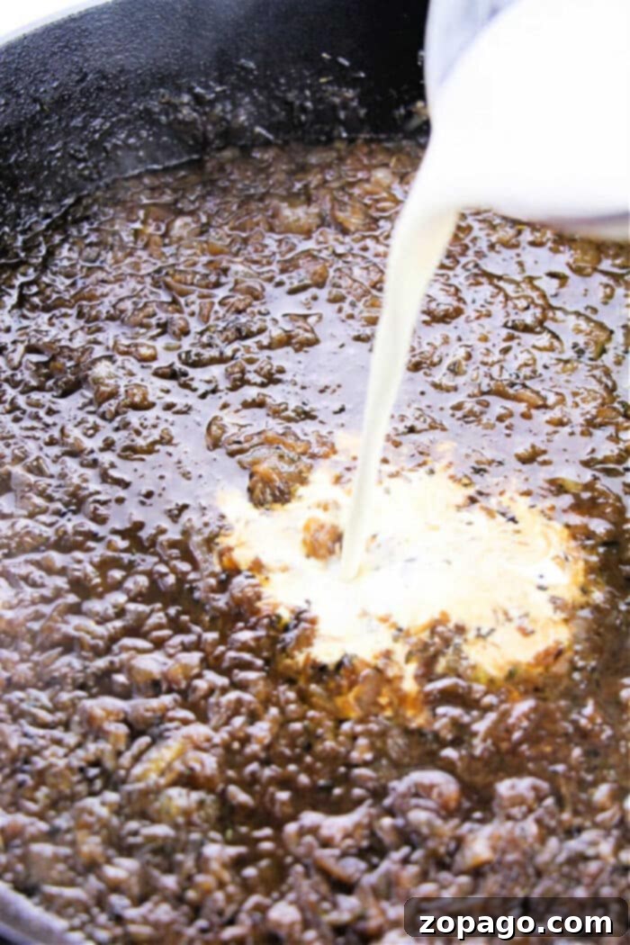 Savory Onion Gravy 4 A stream of rich, heavy whipping cream being poured into a skillet of beautifully browned ingredients, creating a luxurious, creamy base. This step is crucial for achieving the perfect velvety texture in our easy caramelized onion gravy recipe.